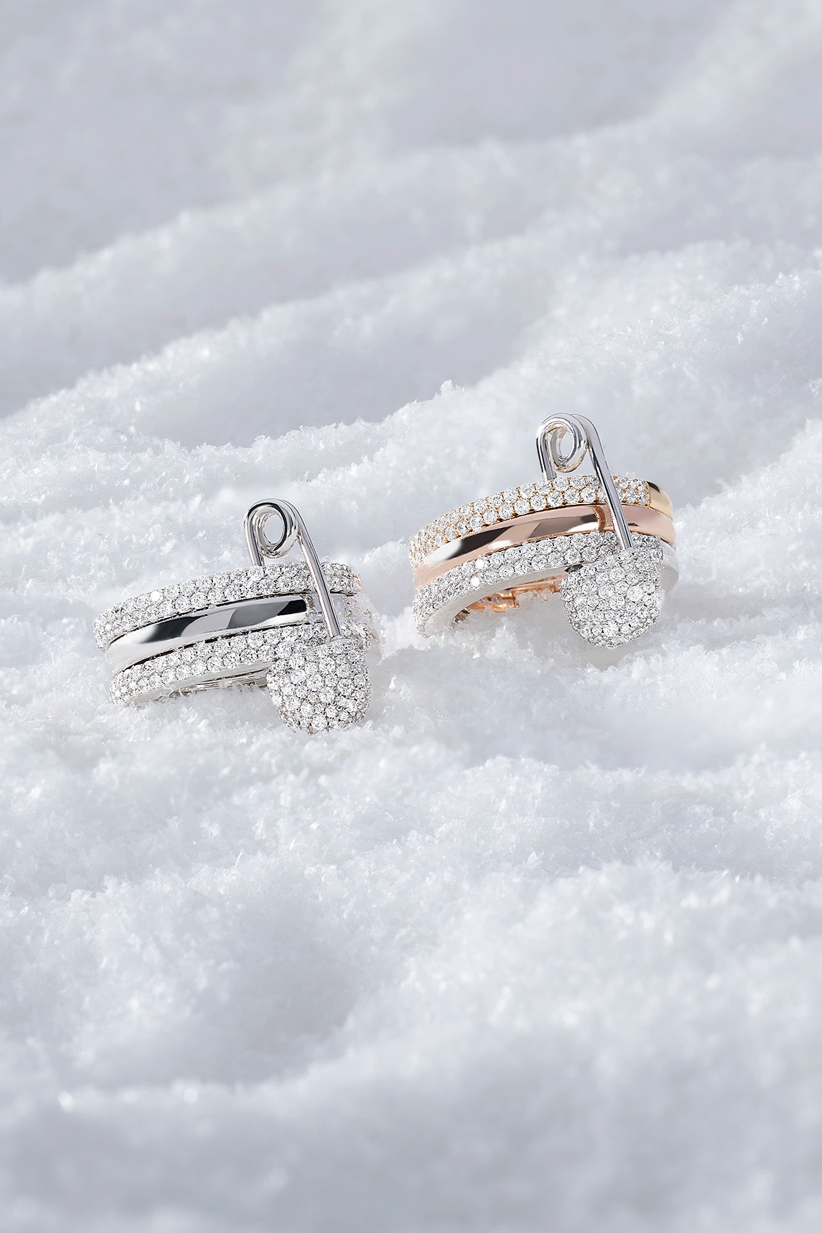 Two rings, one silver and one rose gold, with triple hoop safety pin design from APM MONACO, on a snowy surface.