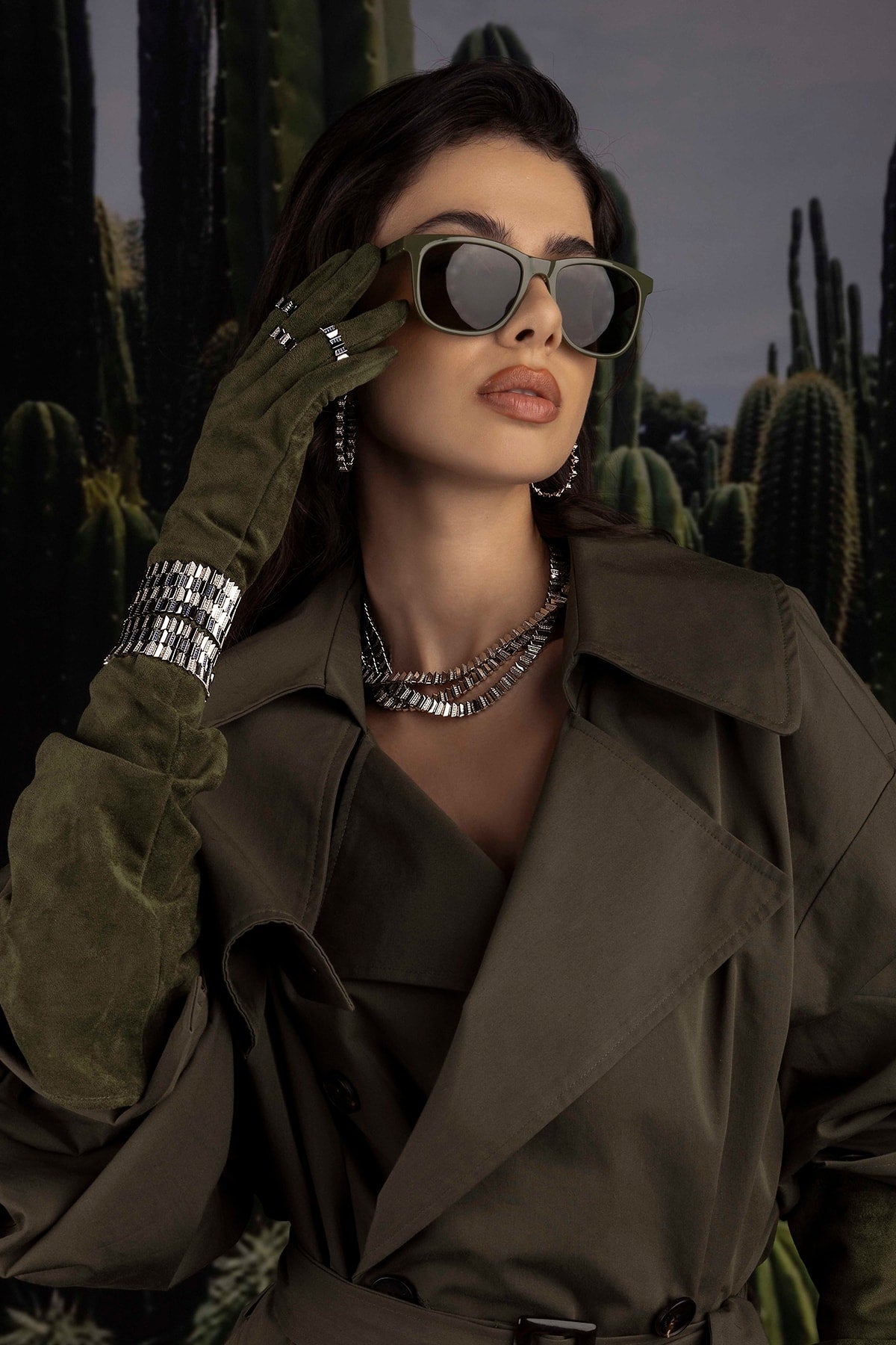 Model in a trench coat, sunglasses and jewelry with cacti in the background