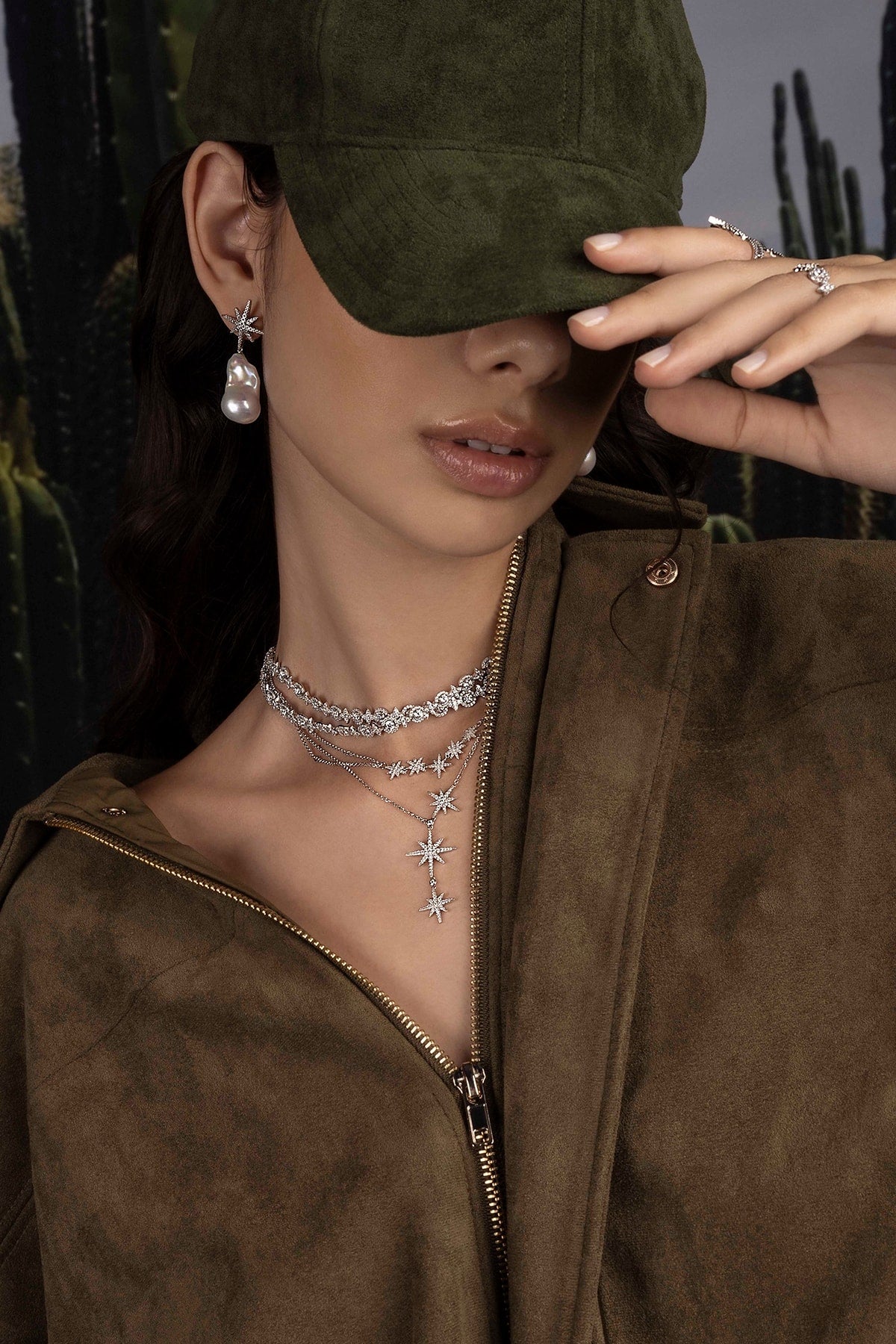 Woman wearing a green cap and brown jacket with layered necklaces against a blurred natural background