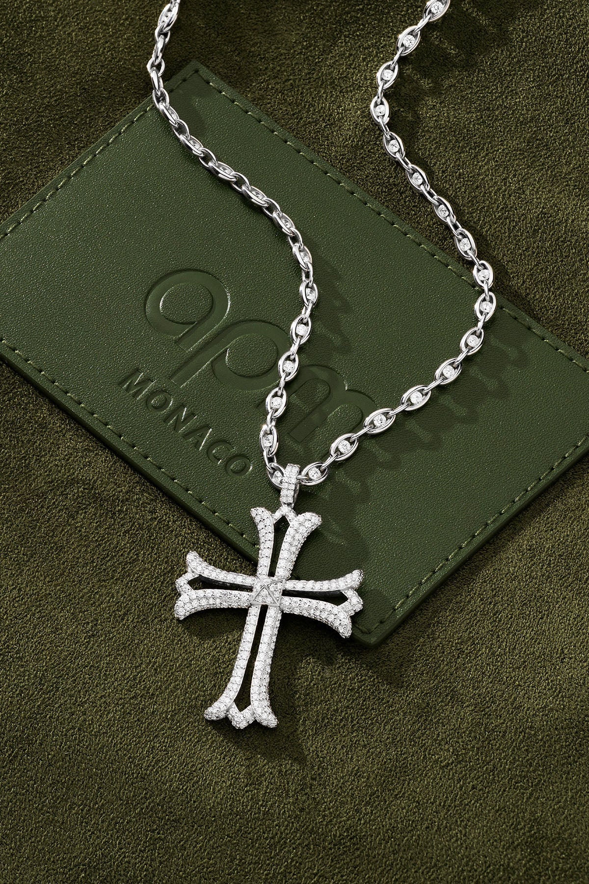 Silver cross necklace on a green textured surface with a brand logo.