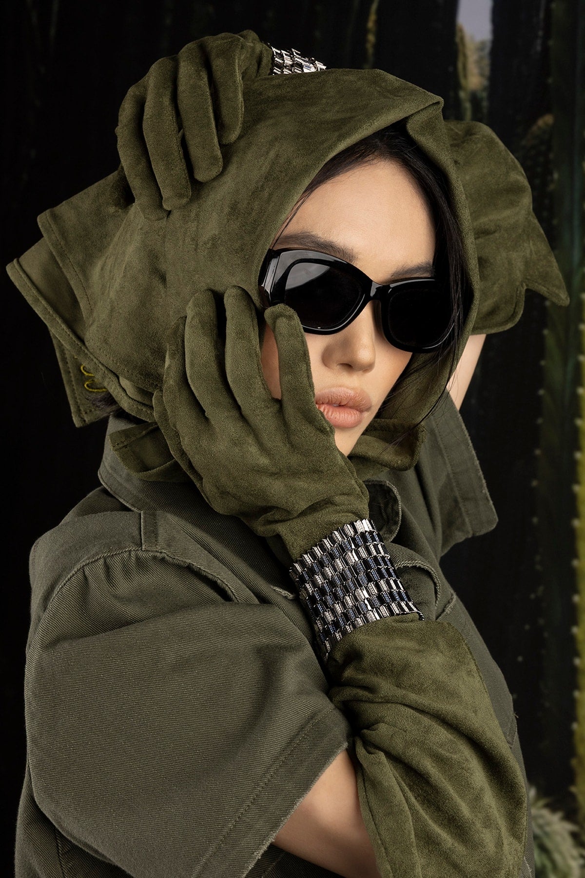 Model wearing a green hooded outfit with gloves and sunglasses against a dark background