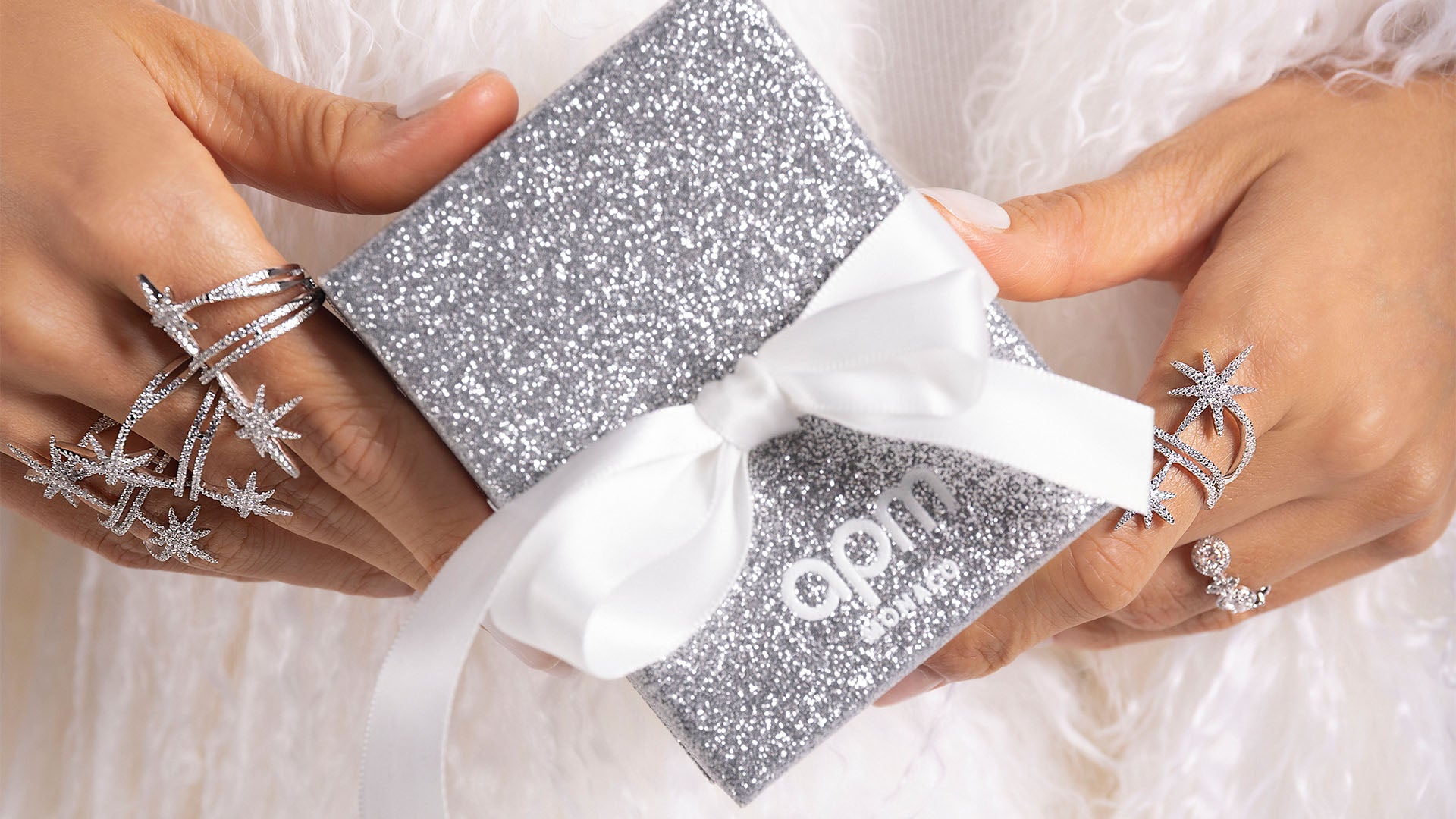 Silver glittery gift box with white ribbon held by hands, featuring the brand 'APM'.