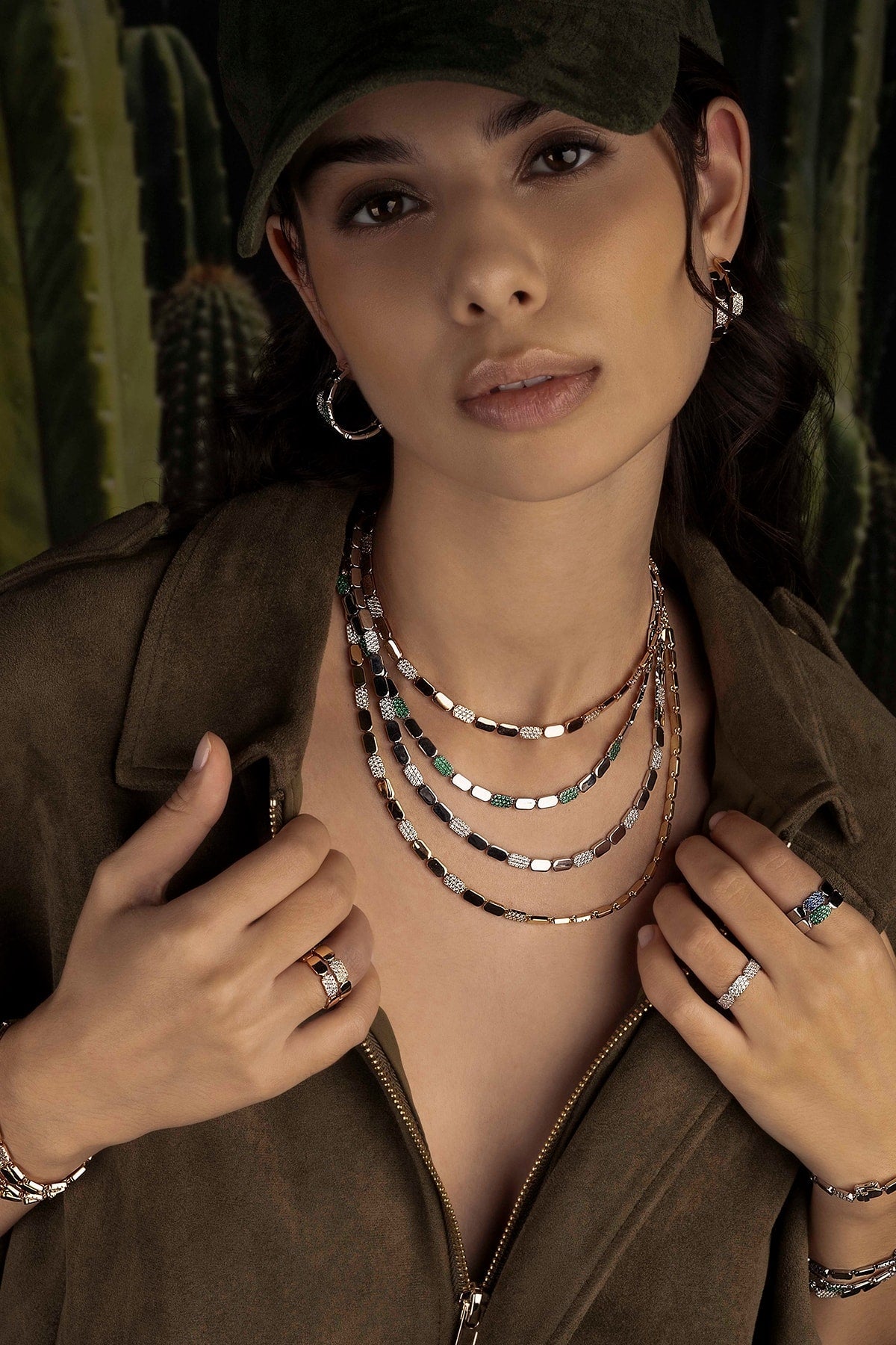 Woman wearing layered necklaces, a brown jacket, and multiple rings with a cactus background
