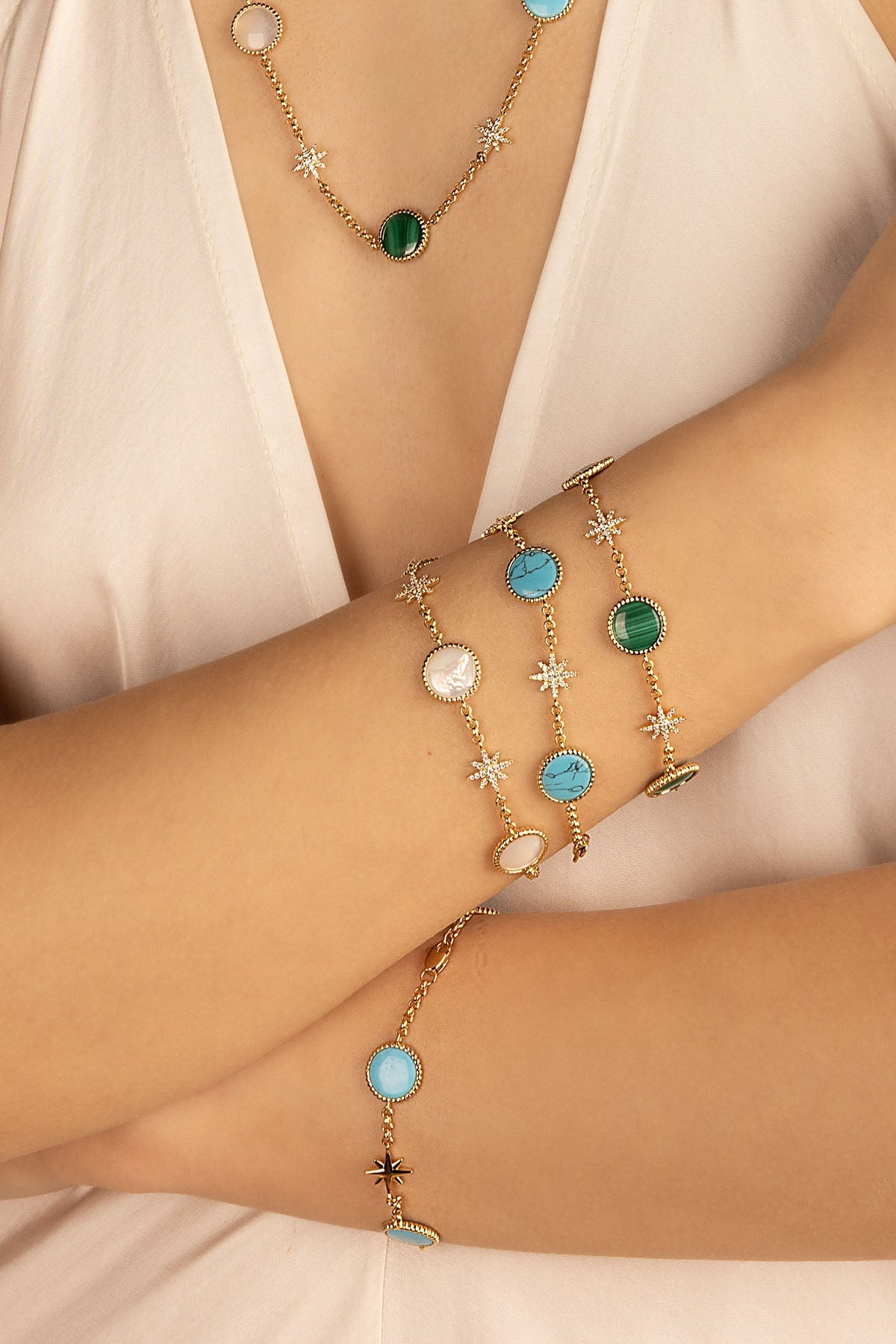 Bracelets APM Monaco. Gold charm bracelet set featuring round blue, green, and white stones with star-shaped crystal accents; paired with a matching charm necklace visible on neckline.