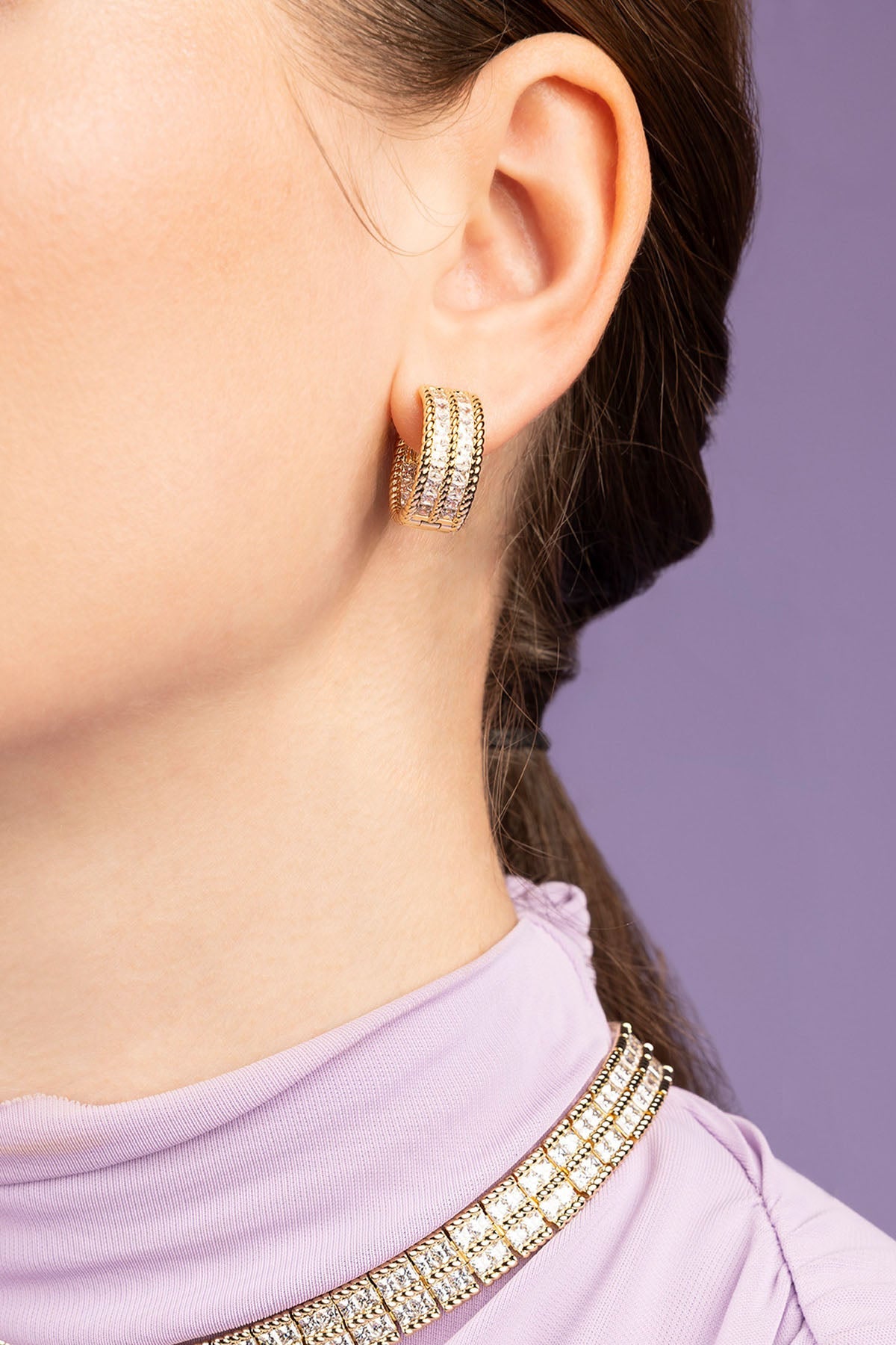 Close-up of a person wearing gold hoop earrings with a blurred purple background