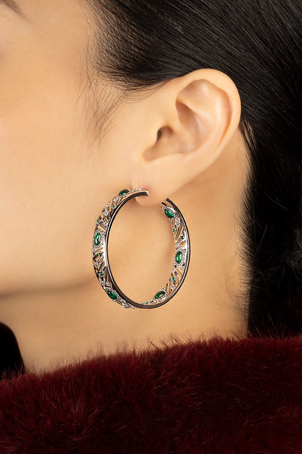 Woman wearing APM hoop earrings, emerald green stones glowing with bold elegance.
