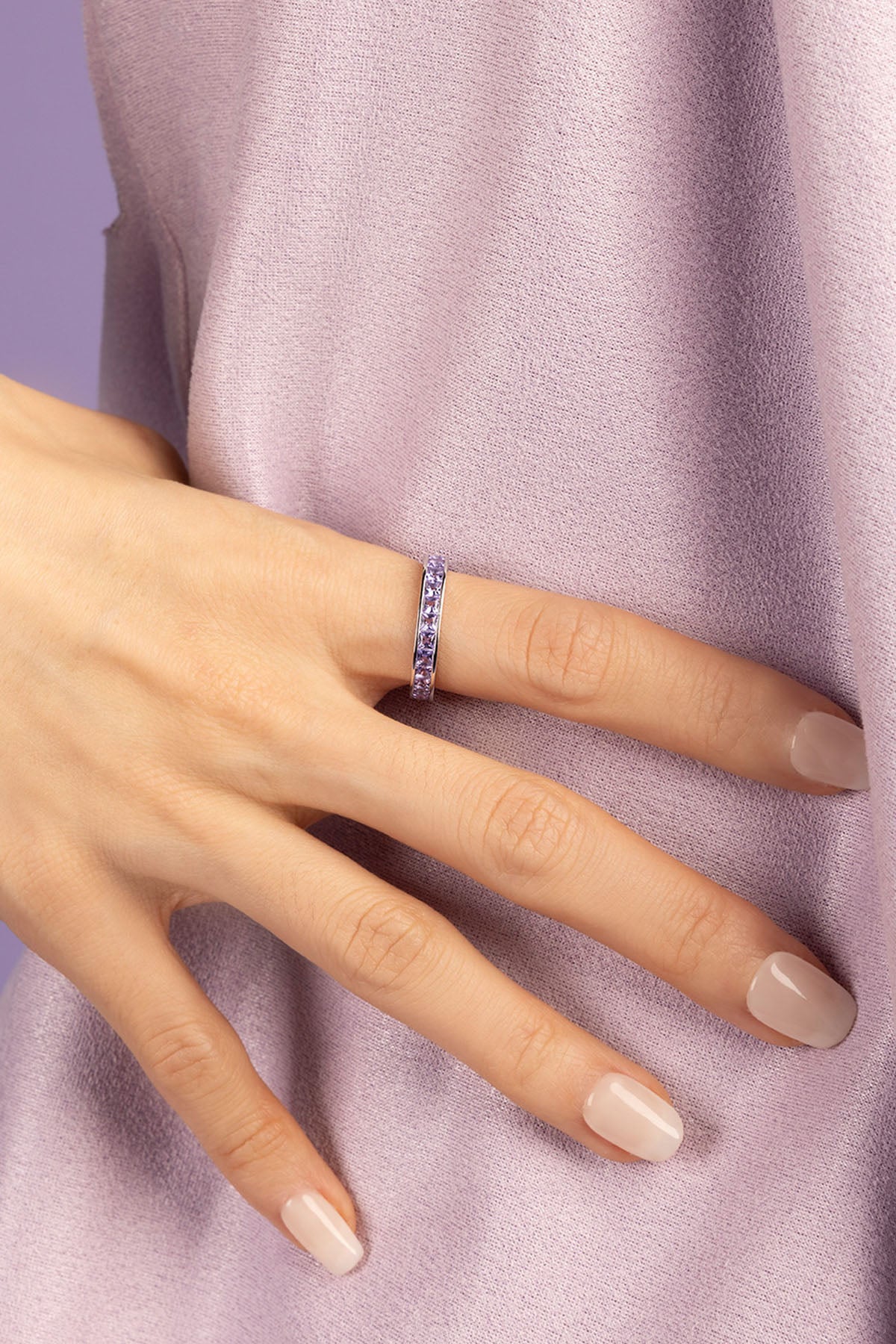 Hand wearing a ring with purple zirconia stones against a light purple background