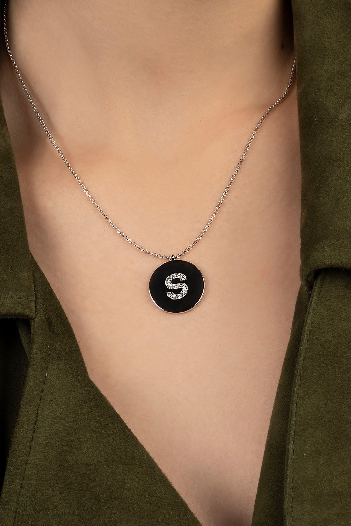 Necklace with a circular pendant featuring the letter 'S' on a person wearing a green shirt.