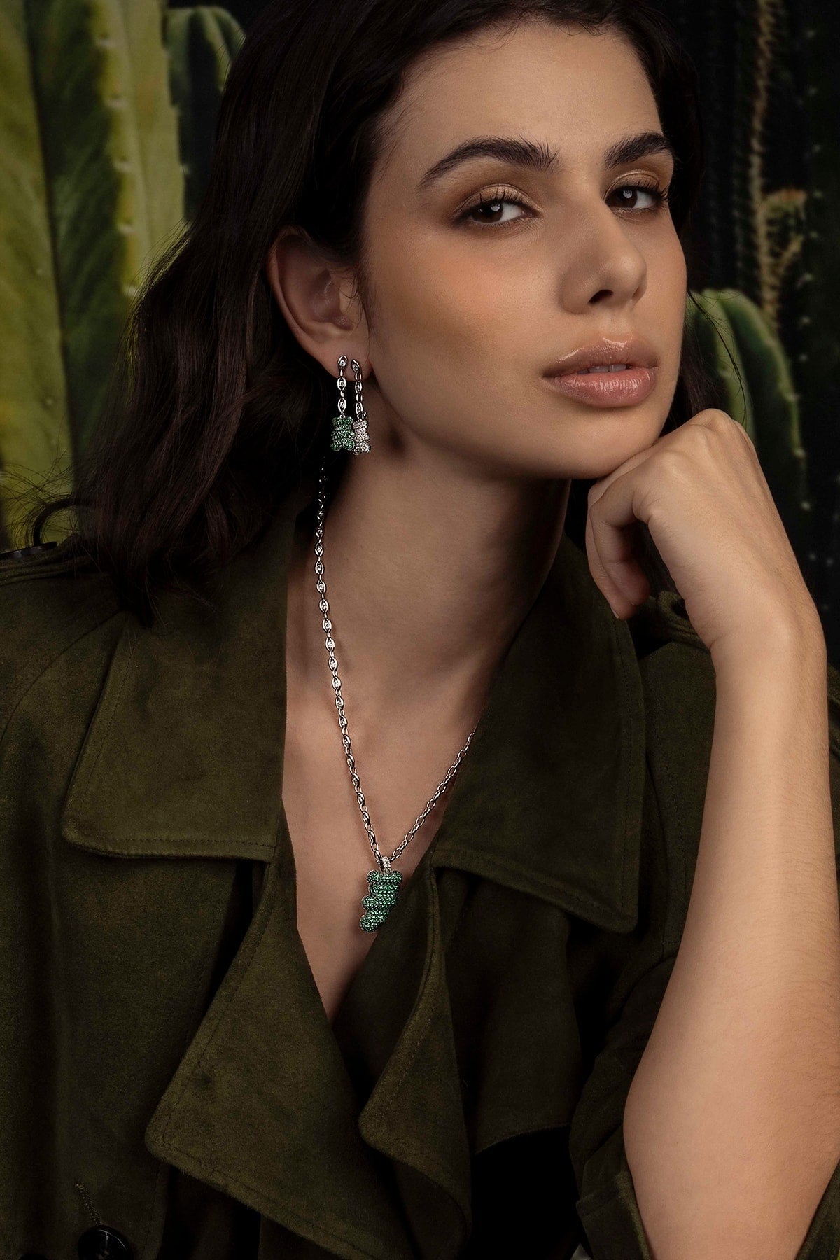 Model wearing a green jacket with jewelry, including earrings and a necklace, against a blurred natural background.