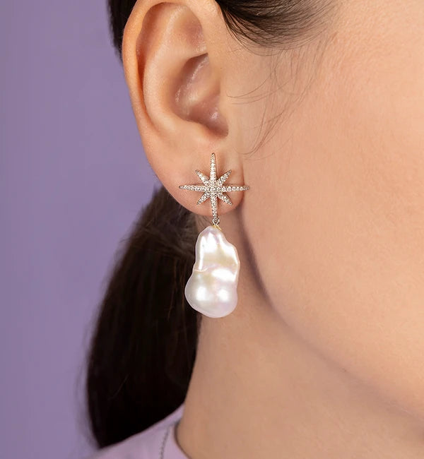 Close-up of an ear wearing a star-shaped APM MONACO earring with a pearl drop against a purple background