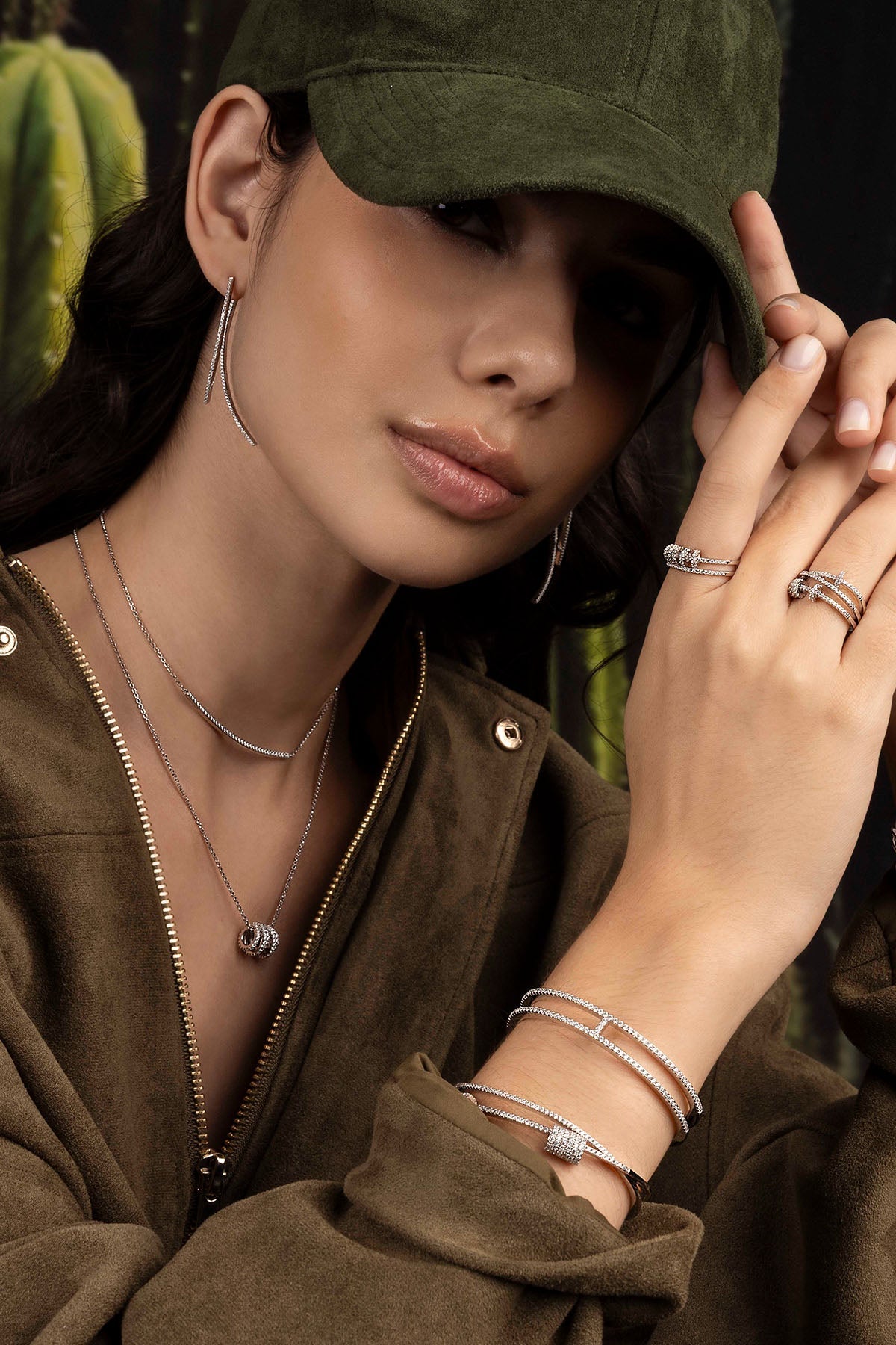 Woman wearing a green cap and brown jacket with jewelry, surrounded by greenery.