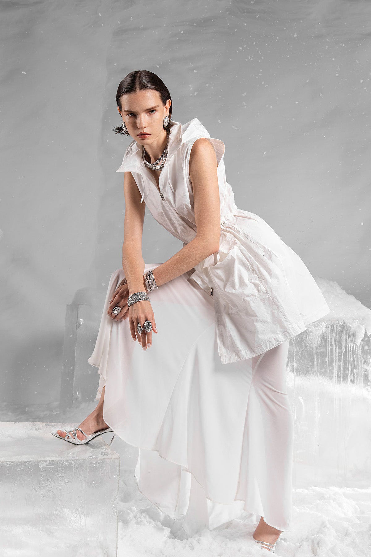 Model in a white dress with APM MONACO whole labyrinth set jewelry sitting on a snowy surface with a gray background