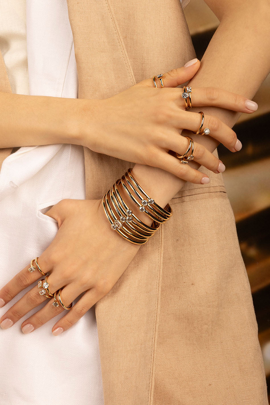 Bracelets APM Monaco and rings in yellow gold with clear stones; cuff-style bracelet on a tan-skinned model, paired with a beige blazer and white shirt.