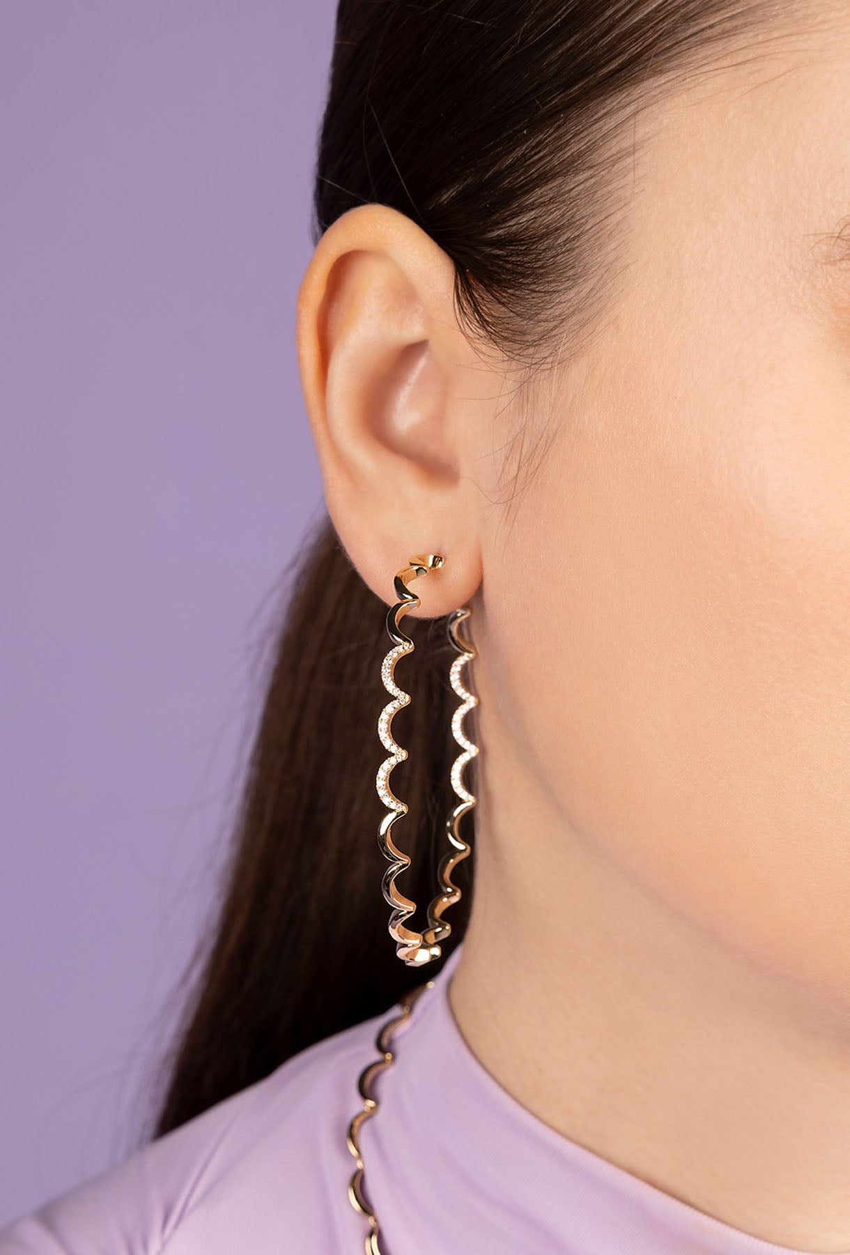Close-up of a person wearing APM MONACO yellow gold wavy hoop earrings with a purple background