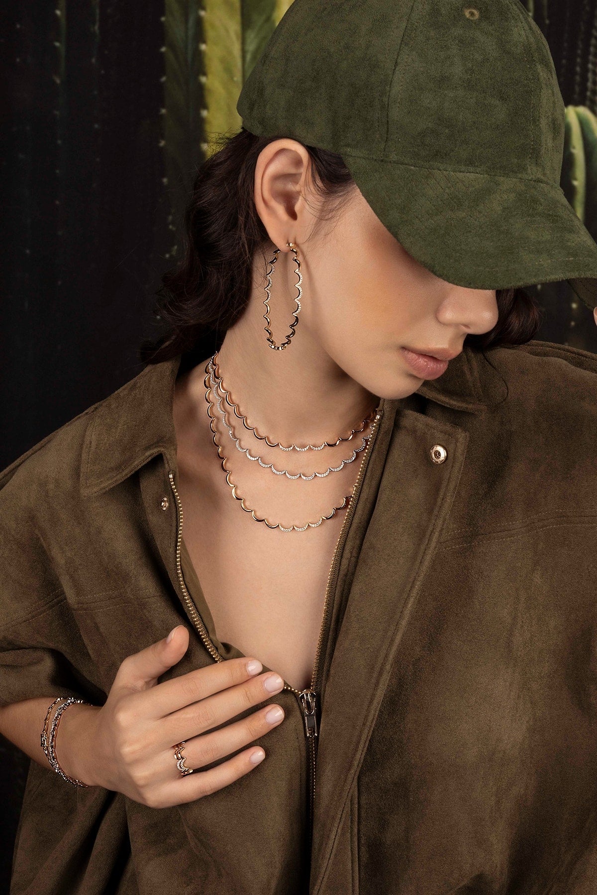 Model wearing a green cap and brown jacket with layered necklaces and a hoop earring against a dark background